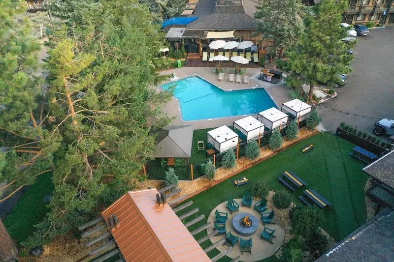Aerial view of Station House Inn, Lake Tahoe with an outdoor swimming pool, fire pit seating area, cabana-style lounges, picnic tables, and pine trees surrounding the property.