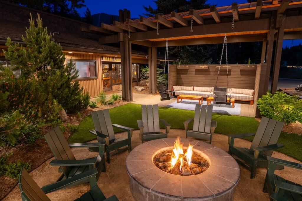 Outdoor fire pit seating area at a mountain lodge, with Adirondack chairs surrounding a stone fire pit, a covered pergola lounge, and warm evening lighting among pine trees.